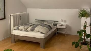 A bedroom with a bed, bedside table, lamp, radiator, potted plant, and a large window.