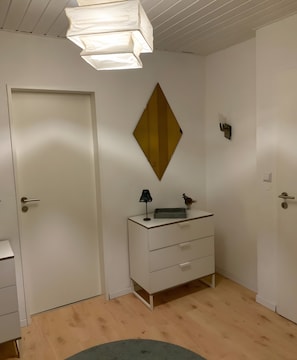 Ein Zimmer mit weißer Tür, einer weißen Kommode mit Schubladen, einem kleinen Tisch, einem wandmontierten Spiegel und einer Deckenleuchte.