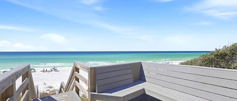 A wooden deck leading to a beach with a bench and a clear view of the ocean.
