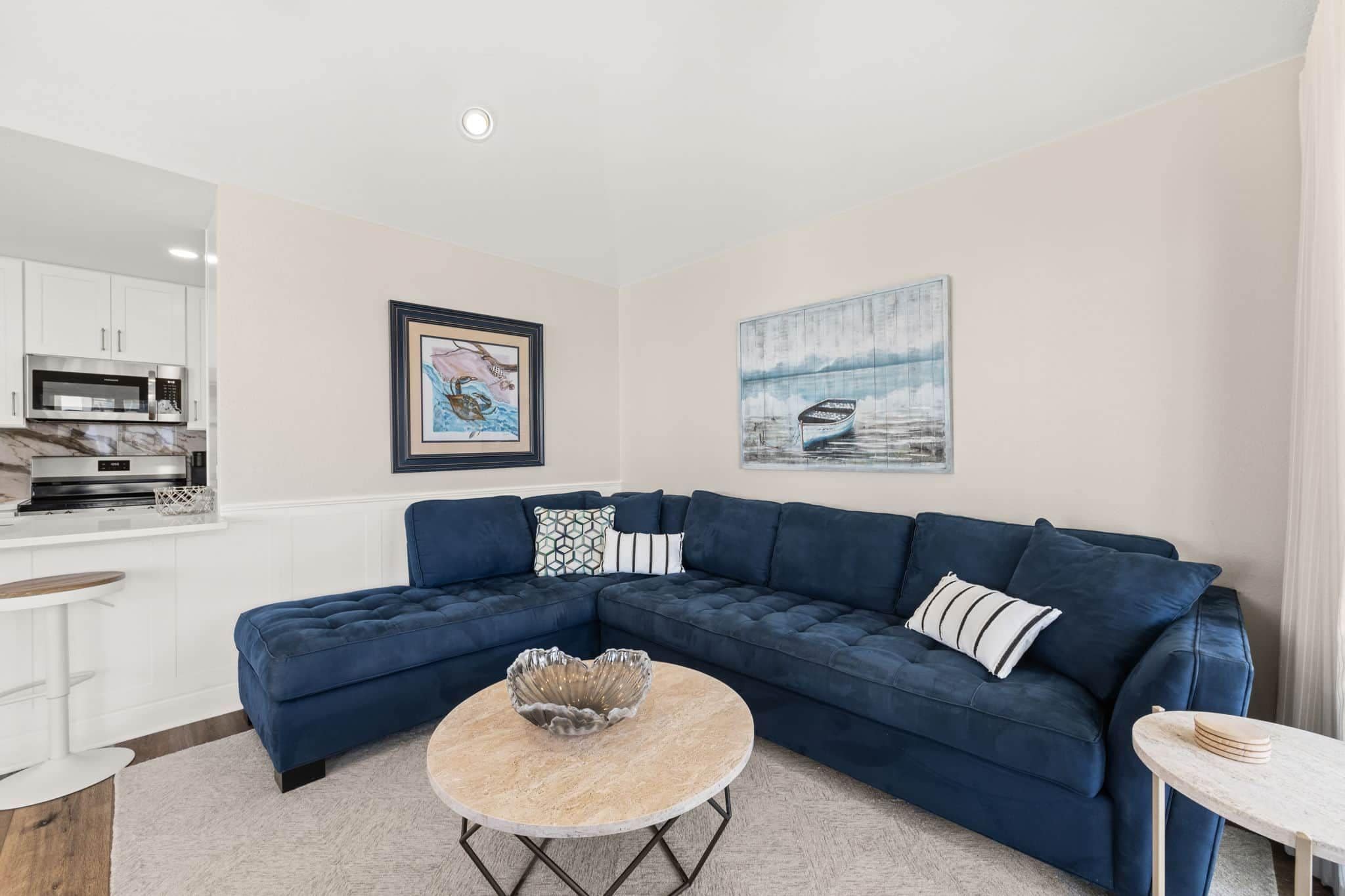 A modern living room with a sectional sofa, a round coffee table, and a kitchen area with white cabinets and a microwave.