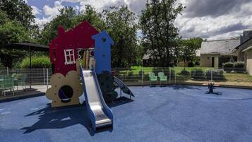 A playground with a slide and climbing structures, surrounded by a fenced area with chairs and trees.