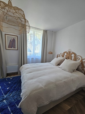 A bedroom with a large bed, a hanging light fixture, a radiator, and a window with curtains. - Room 102 at Villa du Clos Normand, 7 minutes' walk from the sea! (Saint-Martin-aux-Buneaux)