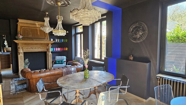 A modern living room with a round dining table, clear chairs, a leather sofa, and a chandelier.