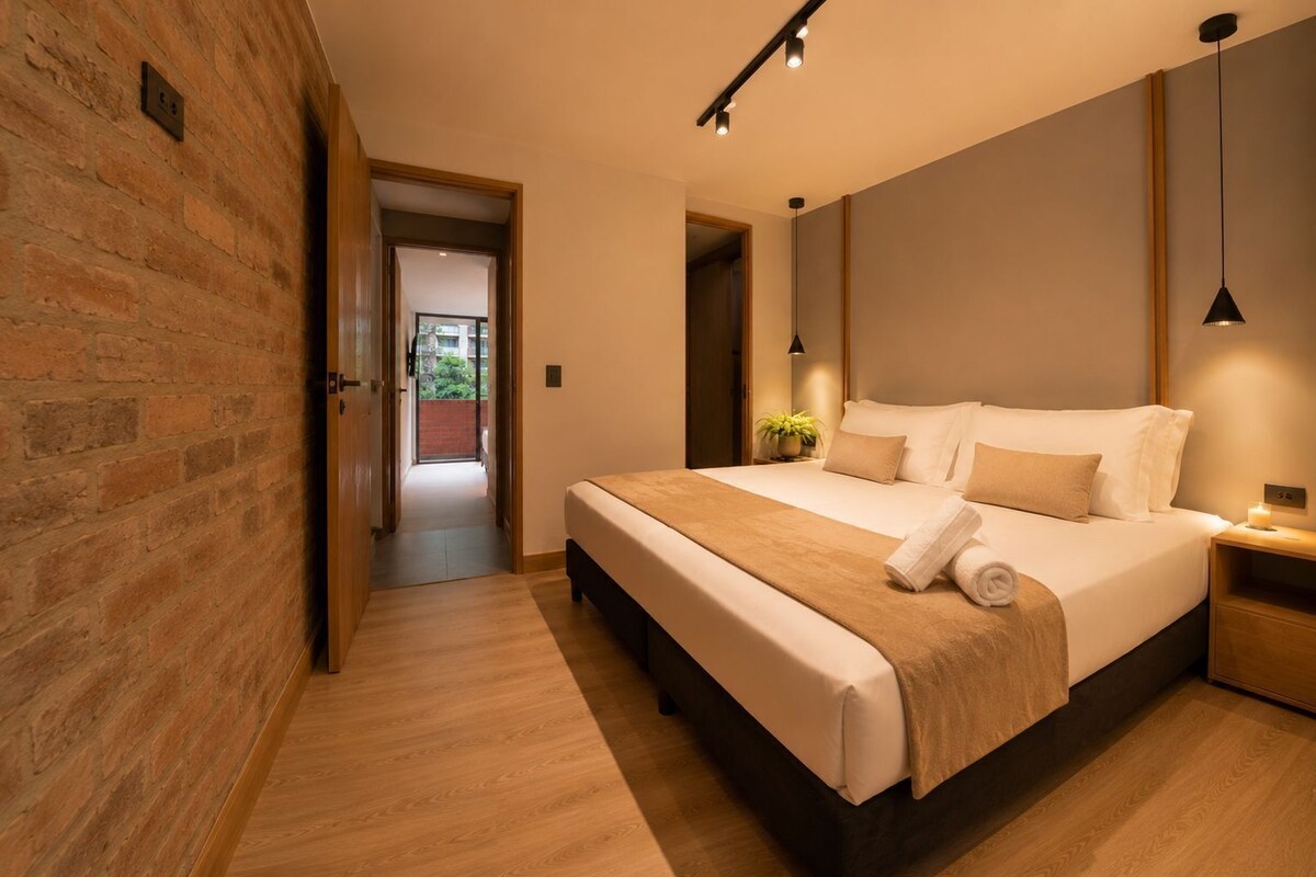 A modern bedroom with a large bed, wooden flooring, a brick wall, and a view through an open door.