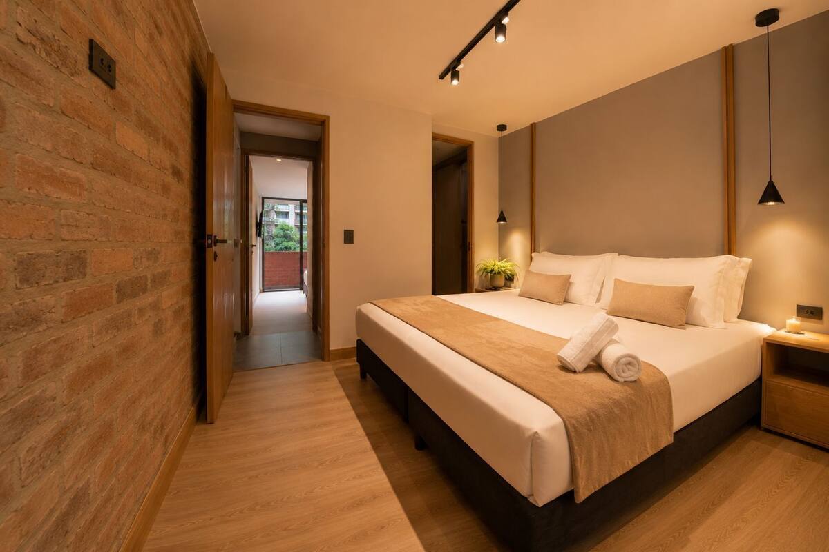 Una camera da letto moderna con un letto grande, pavimento in legno, parete in mattoni e vista attraverso una porta aperta.