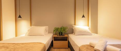 Habitación de hotel con dos camas, una mesita de noche con una planta e iluminación colgante.