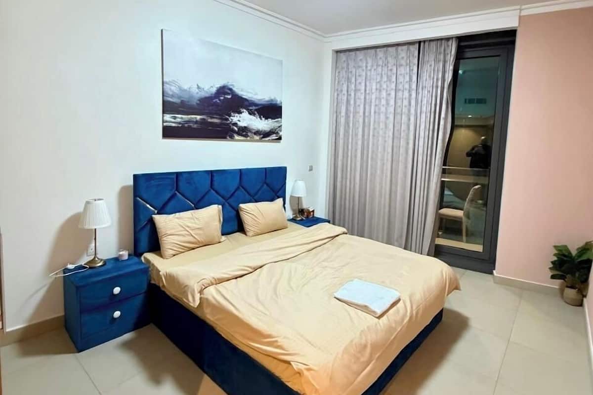Un dormitorio con cama, mesitas de noche, un cuadro, una ventana con cortinas y una planta en maceta.