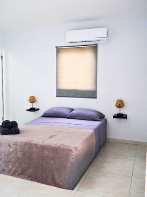 Ein Schlafzimmer mit Bett, Kissen, einem Fenster mit Jalousien, einer Klimaanlage und zwei Nachttischlampen.