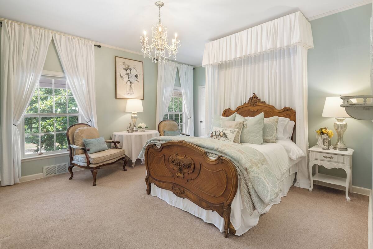 Un dormitorio con una cama grande, una silla, una mesita de noche, un candelabro y cortinas.