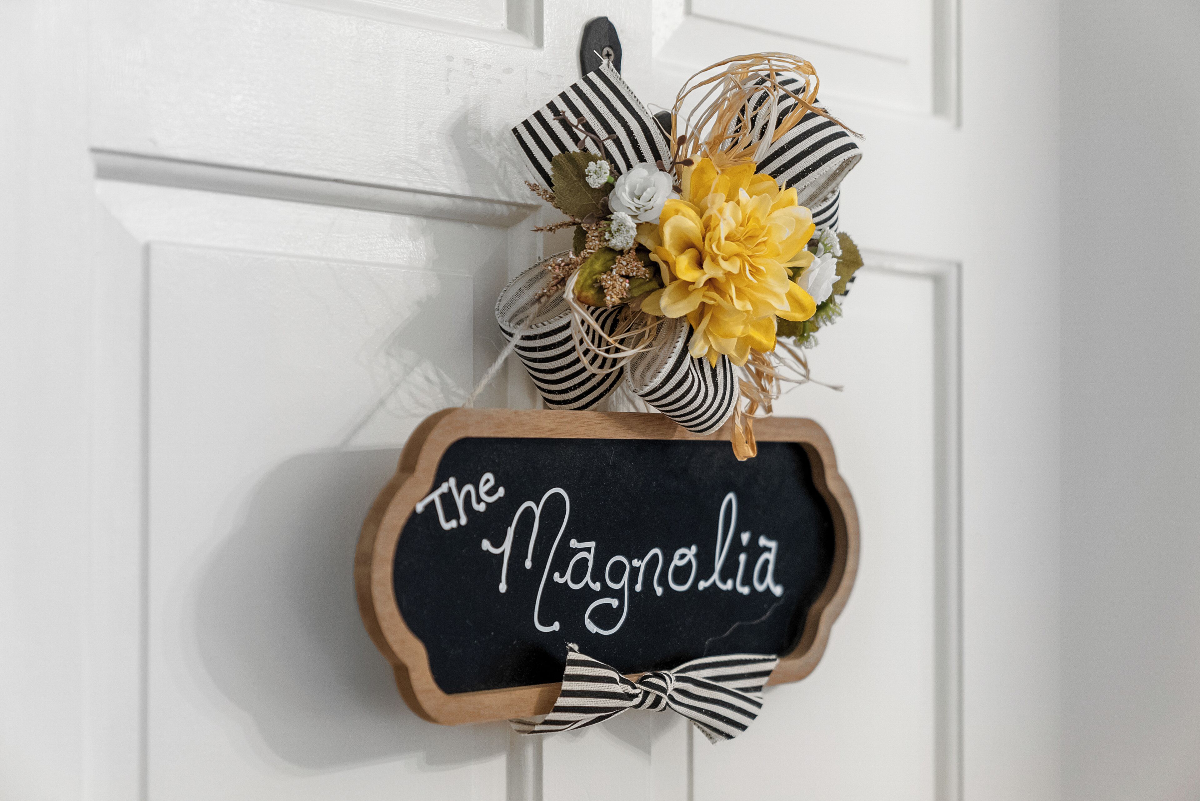 A chalkboard sign with 'The Magnolia' written on it, attached to a white door with a decorative floral arrangement above it.