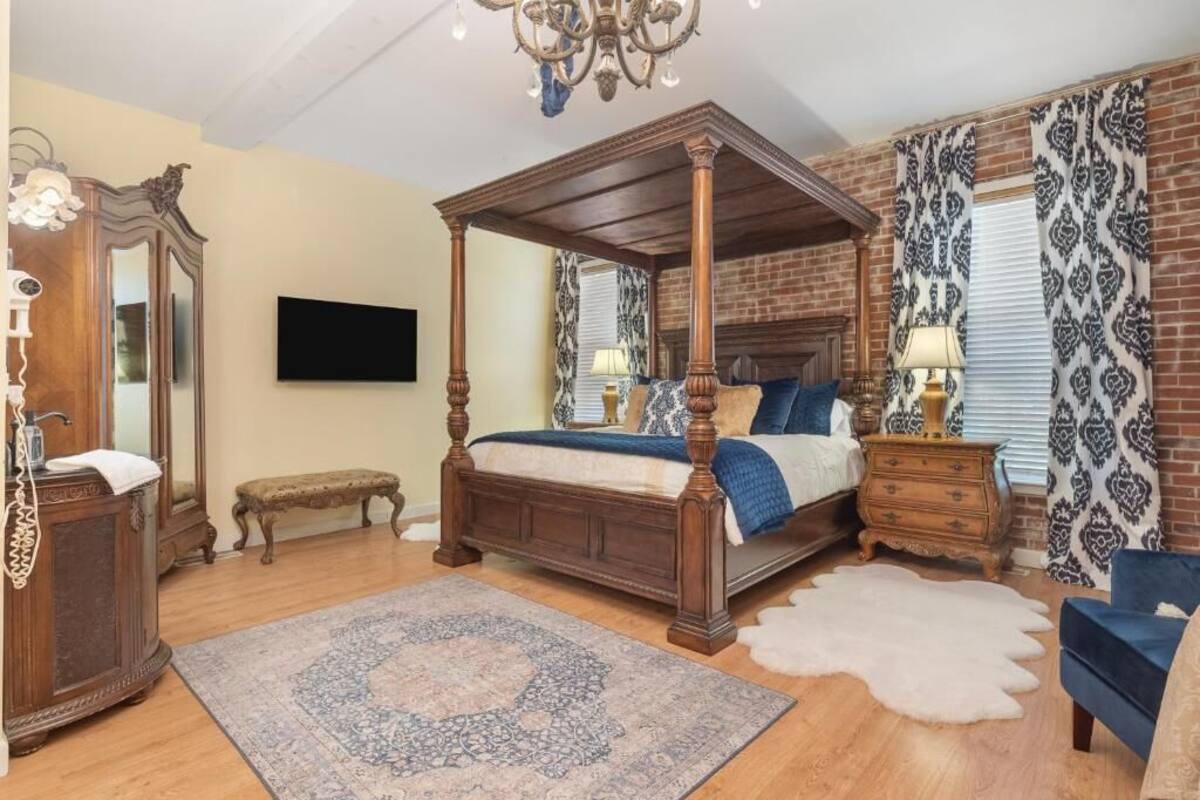Una cama con dosel de madera, un espejo grande, un televisor montado en la pared y una alfombra estampada en el suelo.