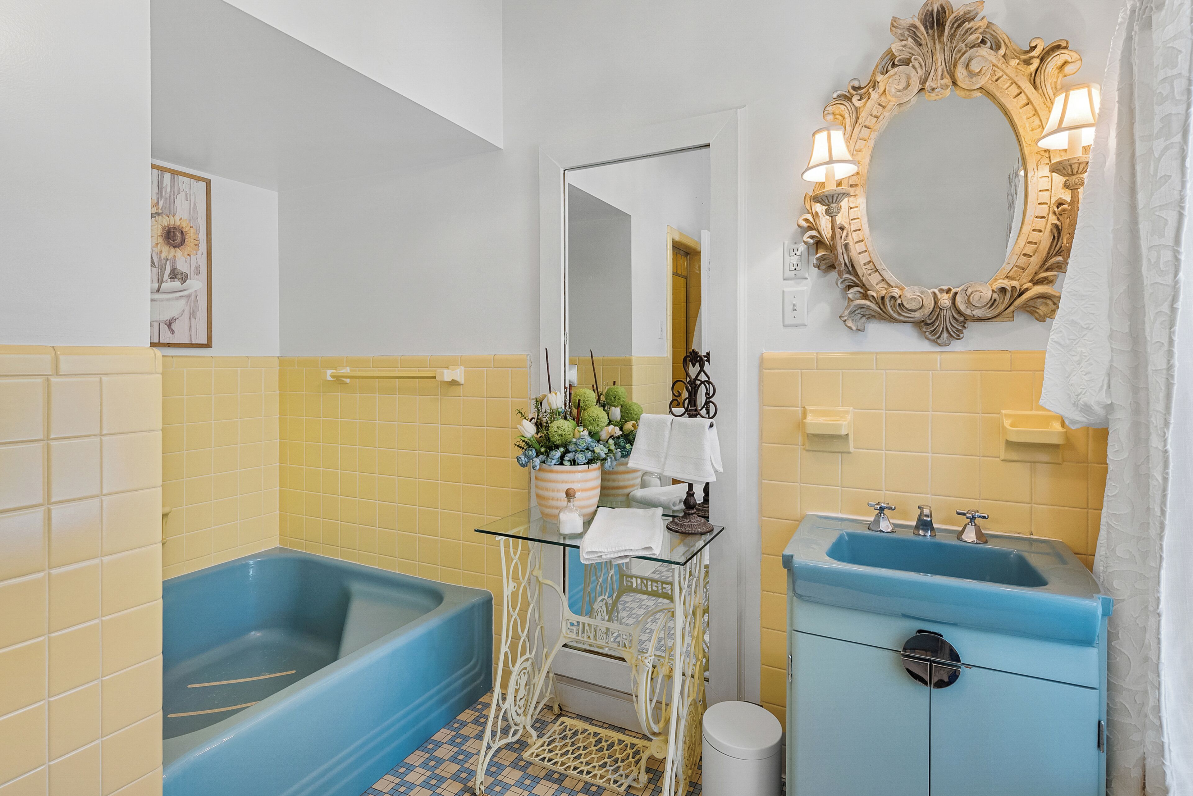 A bathroom with a blue bathtub, a white vanity with a blue sink, a mirror with a decorative frame, and a yellow-tiled wall.