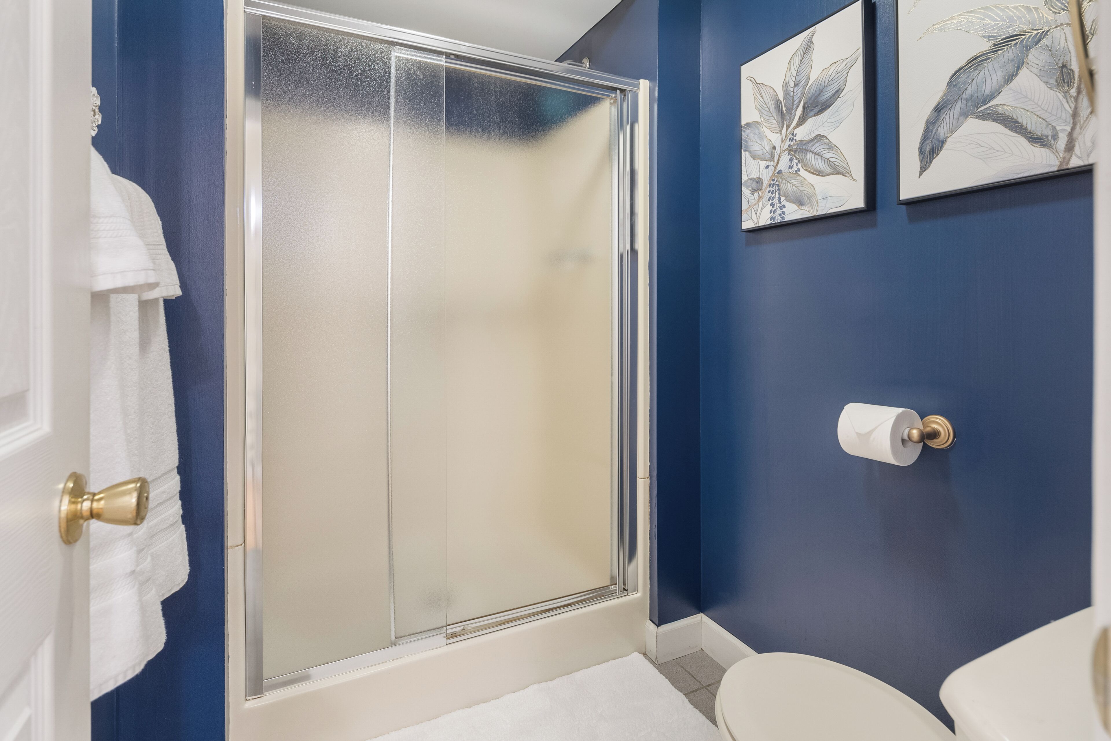 A bathroom with a glass shower door, a toilet, and two framed botanical prints on the wall.