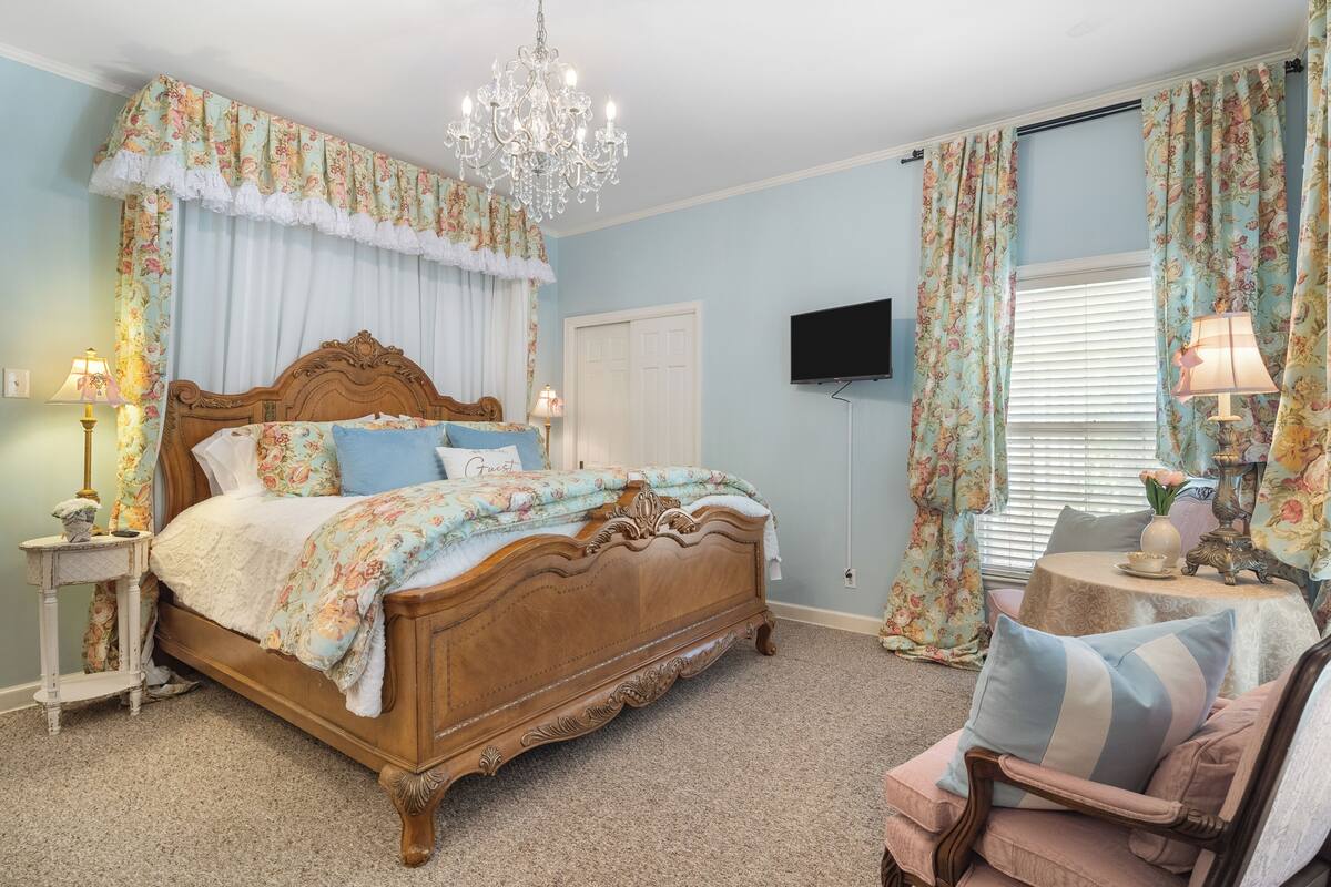 Un dormitorio con cama de madera, candelabro, cortinas con motivos florales, televisor y una mesita de noche con lámpara.