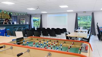 A classroom with a foosball table, chairs, a projector screen, and a window with a view of greenery.