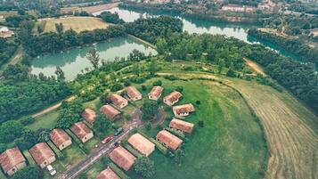 An aerial view of a rural area with a cluster of traditional houses with sloped roofs, surrounded by green fields and a river.