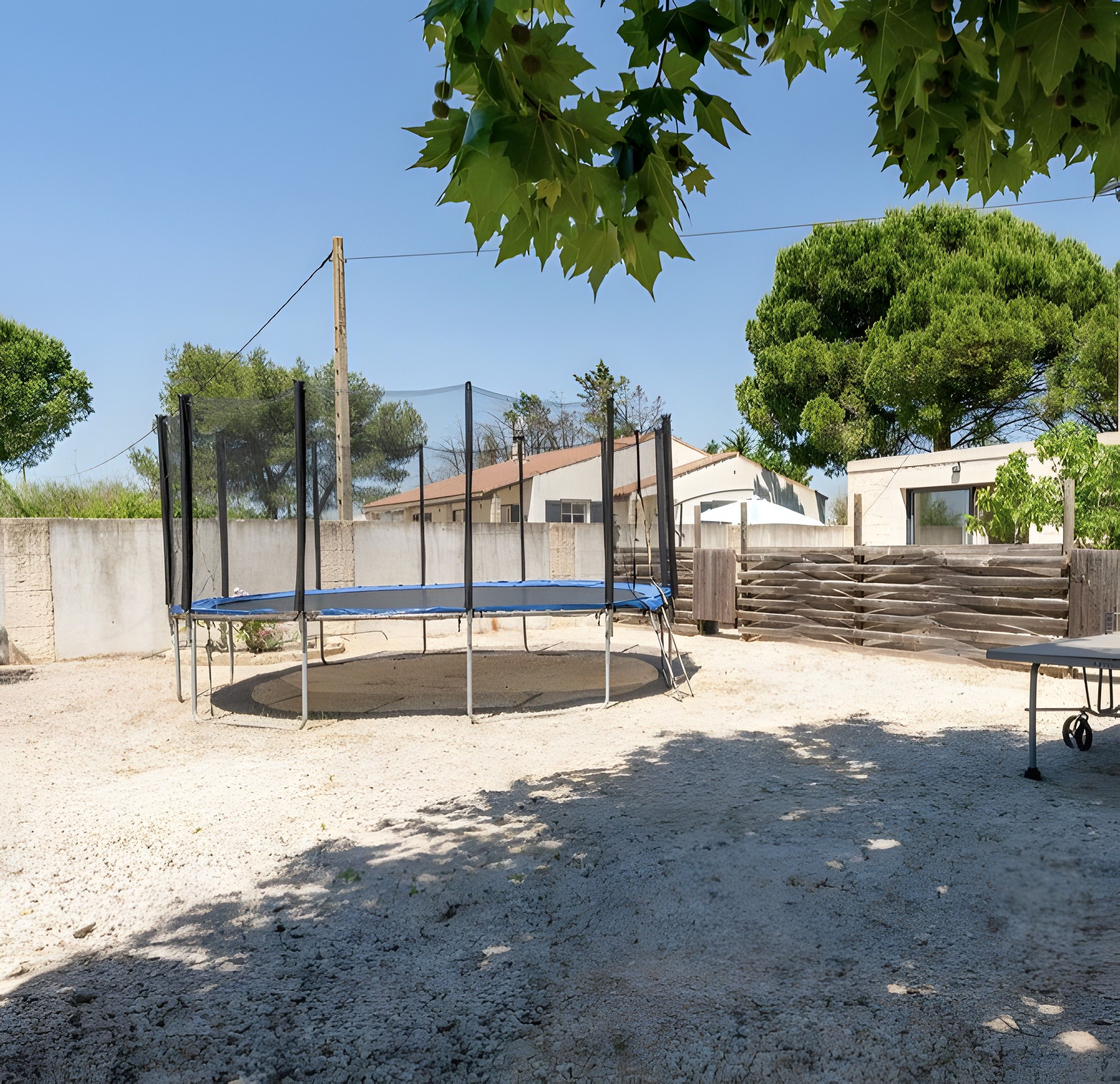 A trampoline in a backyard with a dirt ground, surrounded by a fence, and a building in the background.