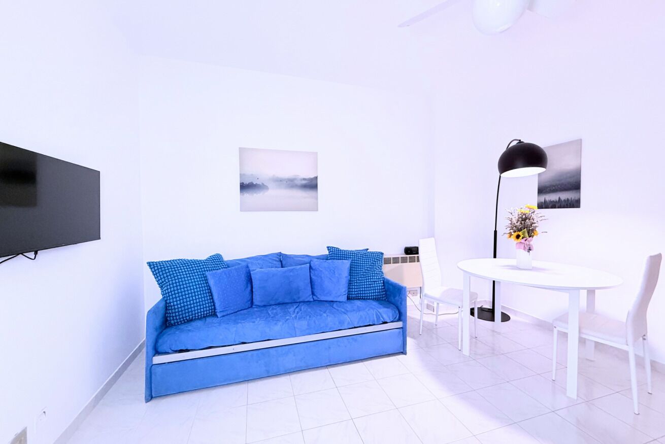 A modern living room with a blue sofa, a white dining table, and a flat-screen TV mounted on the wall.