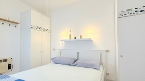 Et soveværelse med seng, hvidt skab, sengeborde med lamper og en lille hylde med pynt.