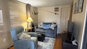 A compact living space with a sofa, a chair, a TV, and a lamp. - “Ocean Breeze” 1BDR, 1Bath cottage on Nettles Island, close to beach and river (Jensen Beach)