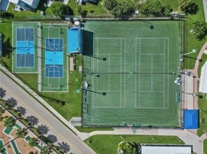 An aerial view of a sports complex with multiple tennis courts, a basketball court, and a large green field.