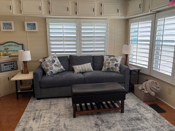 A living room with a grey sofa, two patterned throw pillows, a wooden bench, a small table with a lamp, and a radio.