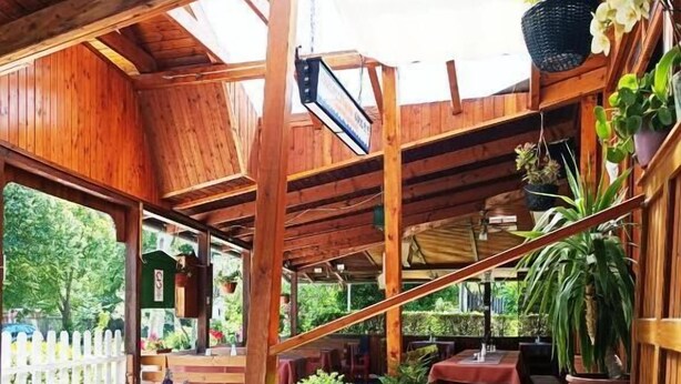 Un espacio exterior de comedor con mesas y bancos de madera, rodeado de plantas en macetas y una cerca de piquetes blancos.