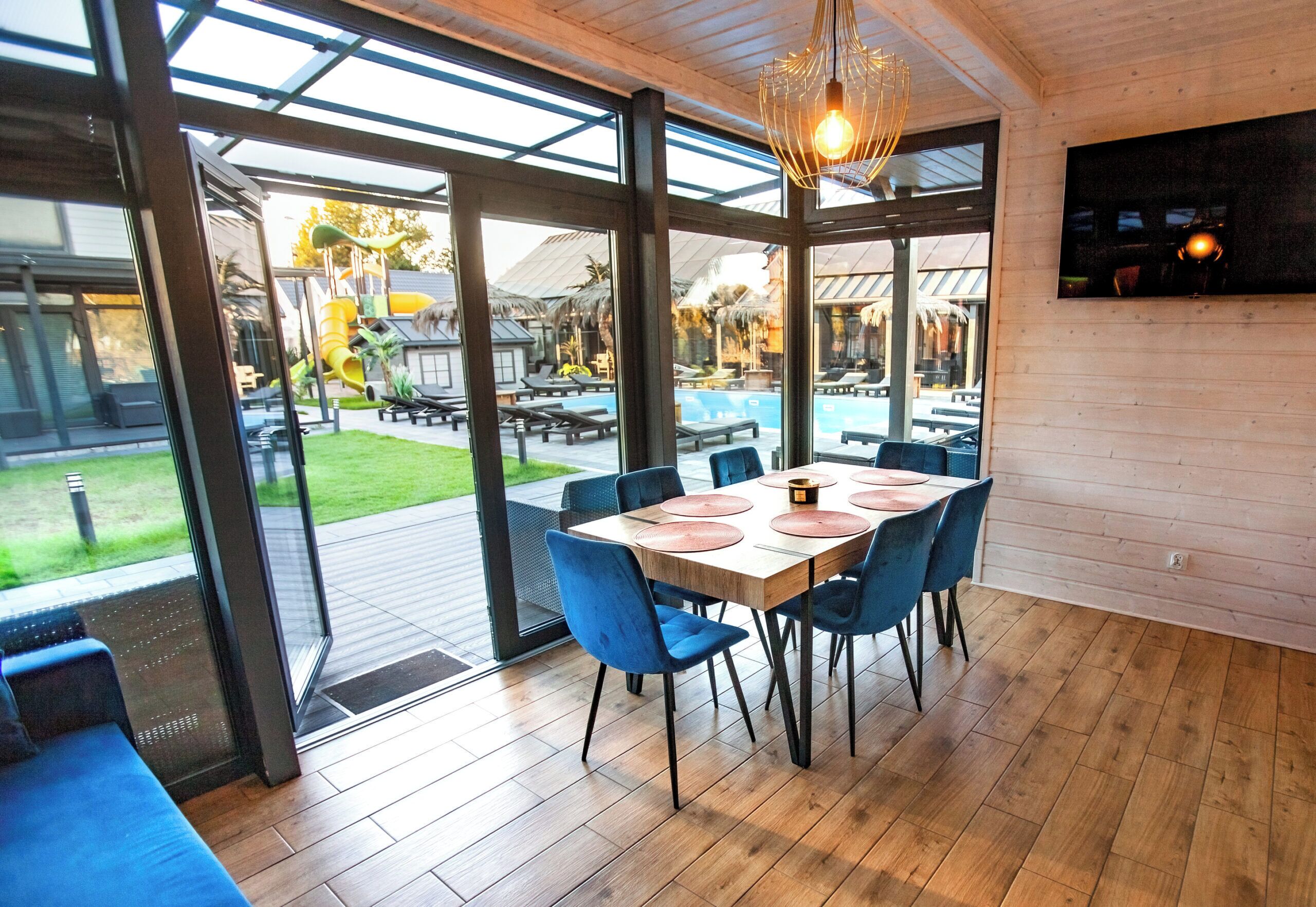 A dining area with a table set for four, surrounded by blue chairs, large windows, and a wooden ceiling.