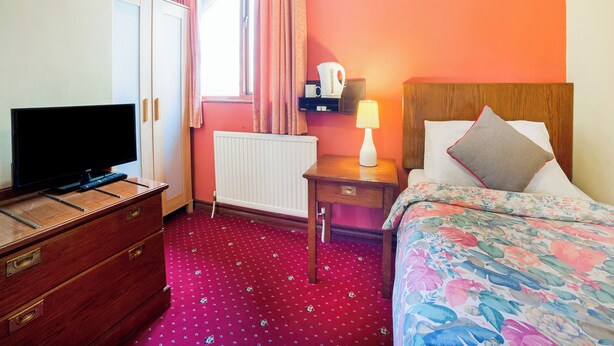 A hotel room with a bed, a TV, a dresser, a nightstand with a lamp, and a window with curtains.