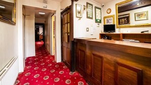 A hotel lobby with a red carpet, wooden reception desk, framed pictures on the wall, and a large mirror.