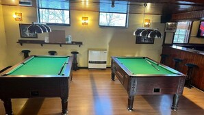 A pool table with a green felt surface, a bar area with stools, and a television mounted on the wall.