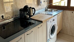 A compact kitchen with wooden cabinets, a built-in stove, a washing machine, and a window with blinds.