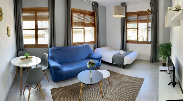 Een moderne kamer met een blauwe bank, een klein rond tafeltje met een plant, een bed met een witte matras en een groot raam met houten jaloezieën.