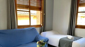 A room with a blue sofa, a small round table with a vase, a bed with a white mattress and a grey blanket, and a hanging lamp.