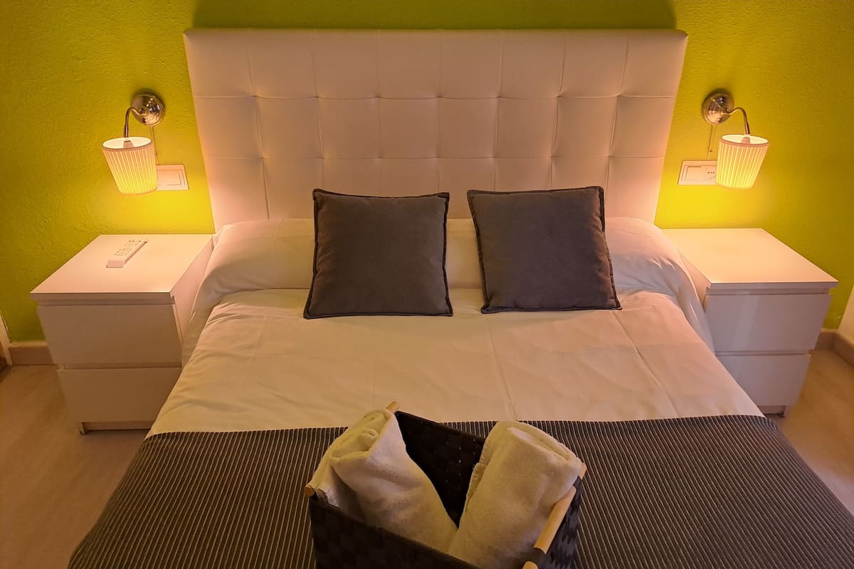 Een netjes opgemaakt bed met twee kussens, een nachtkastje met een lamp en een bijzettafeltje met een afstandsbediening.
