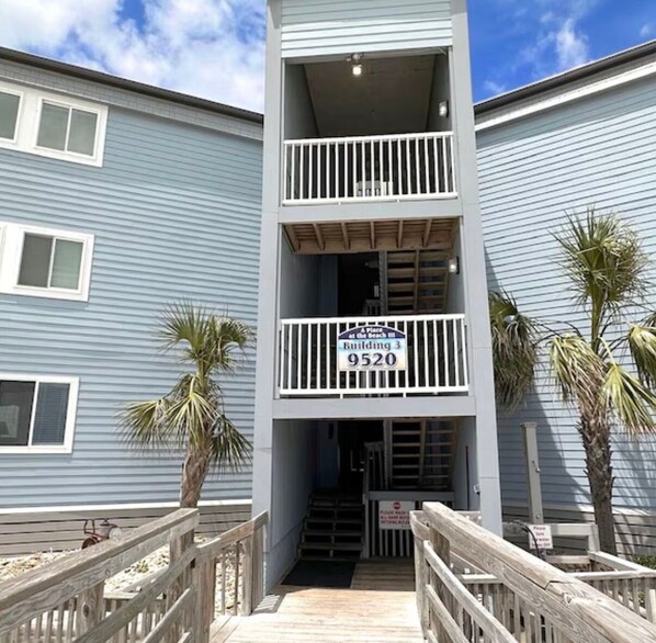 A multi-story building with a balcony on the second floor, a sign indicating 'Building 3 9520,' and a wooden deck leading to the entrance. - Oceanfront•Steps to beach•Pool view•Sleeps 6 (Myrtle beach)
