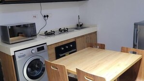 Een kleine keuken met een magnetron, wasmachine en een eethoek met een houten tafel en stoelen.