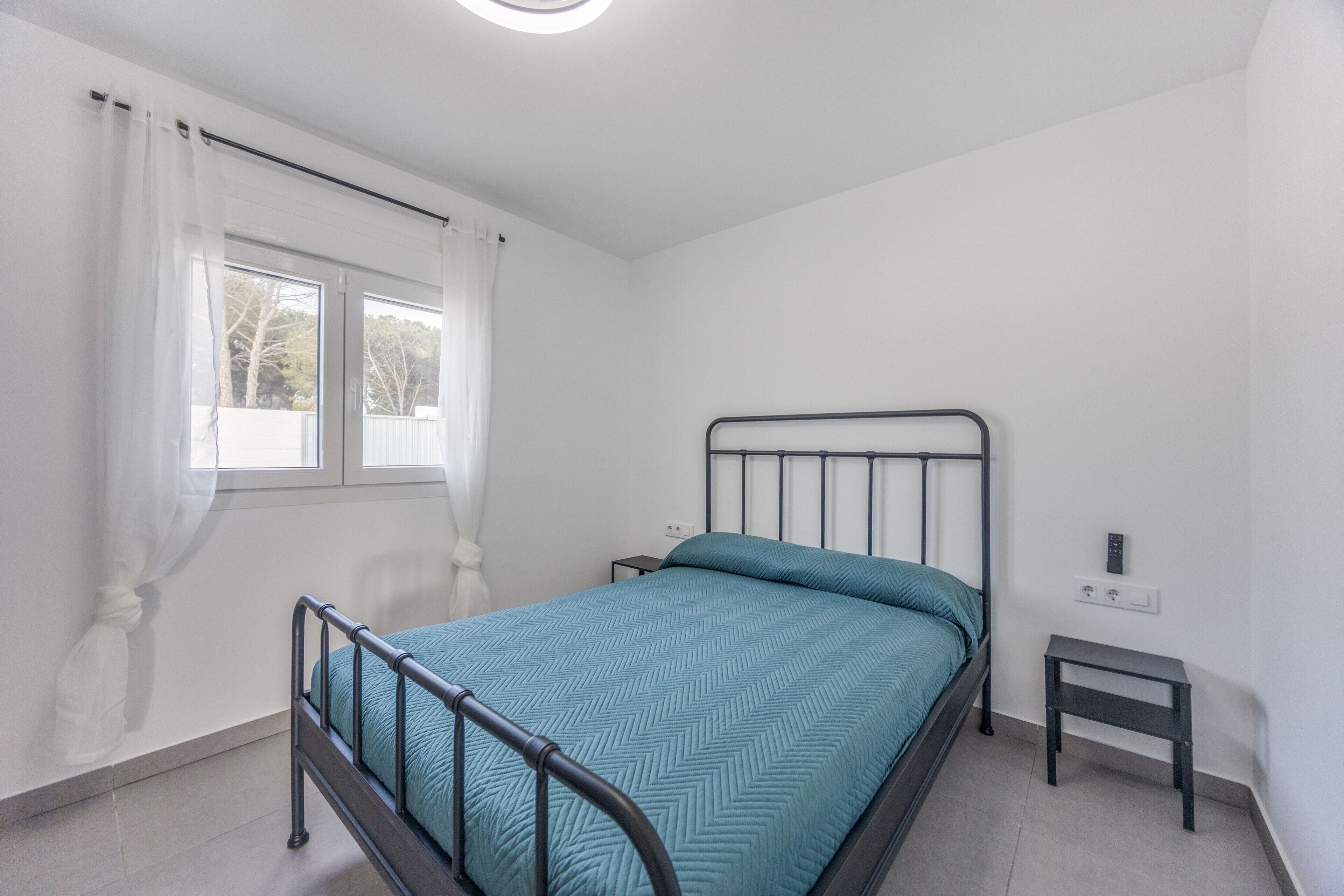 A single bed with a metal frame, a bedside table, a power outlet, and a window with curtains.