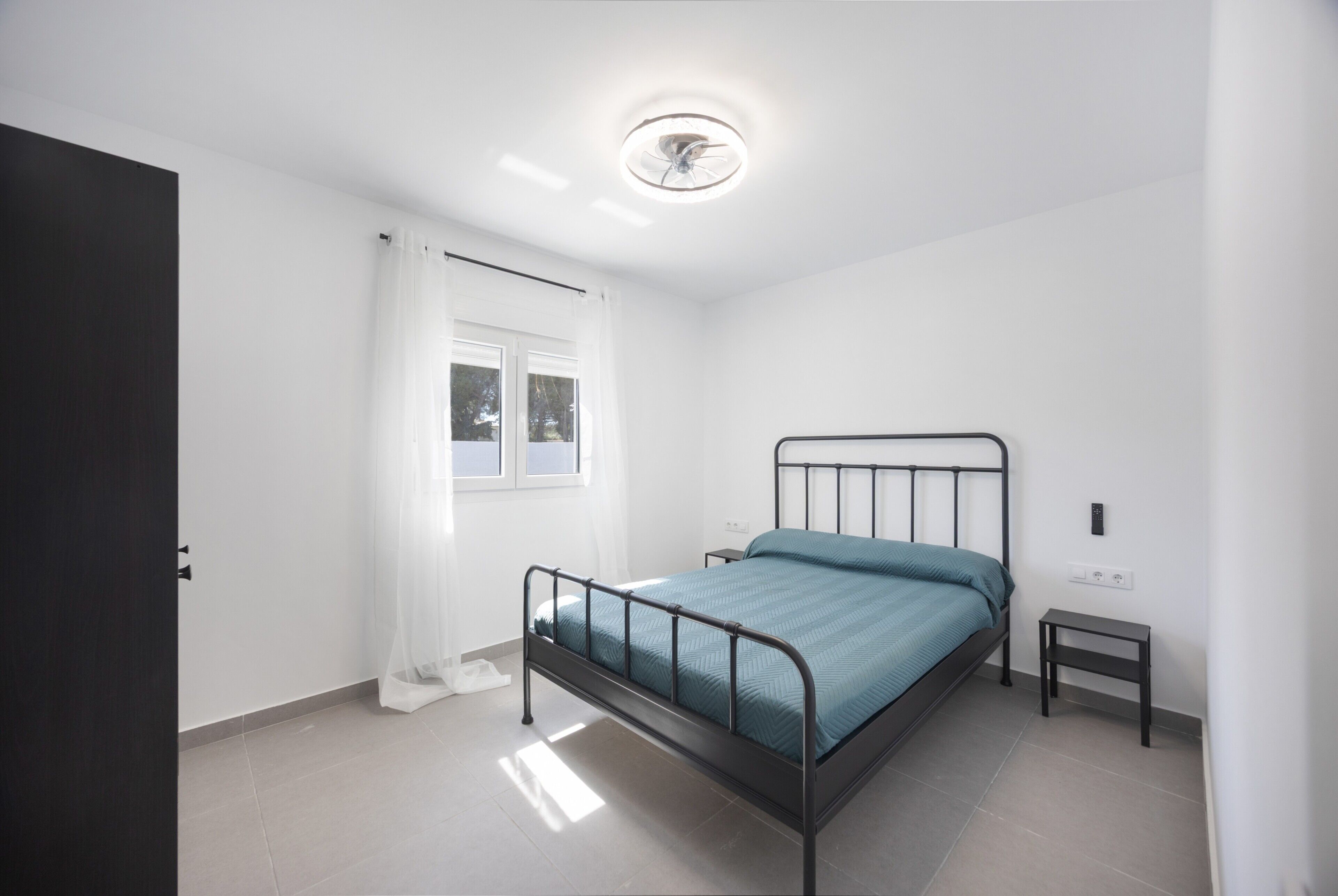 Un dormitorio con cama, mesita de noche, una pequeña mesa, ventana con cortinas e iluminación en el techo.