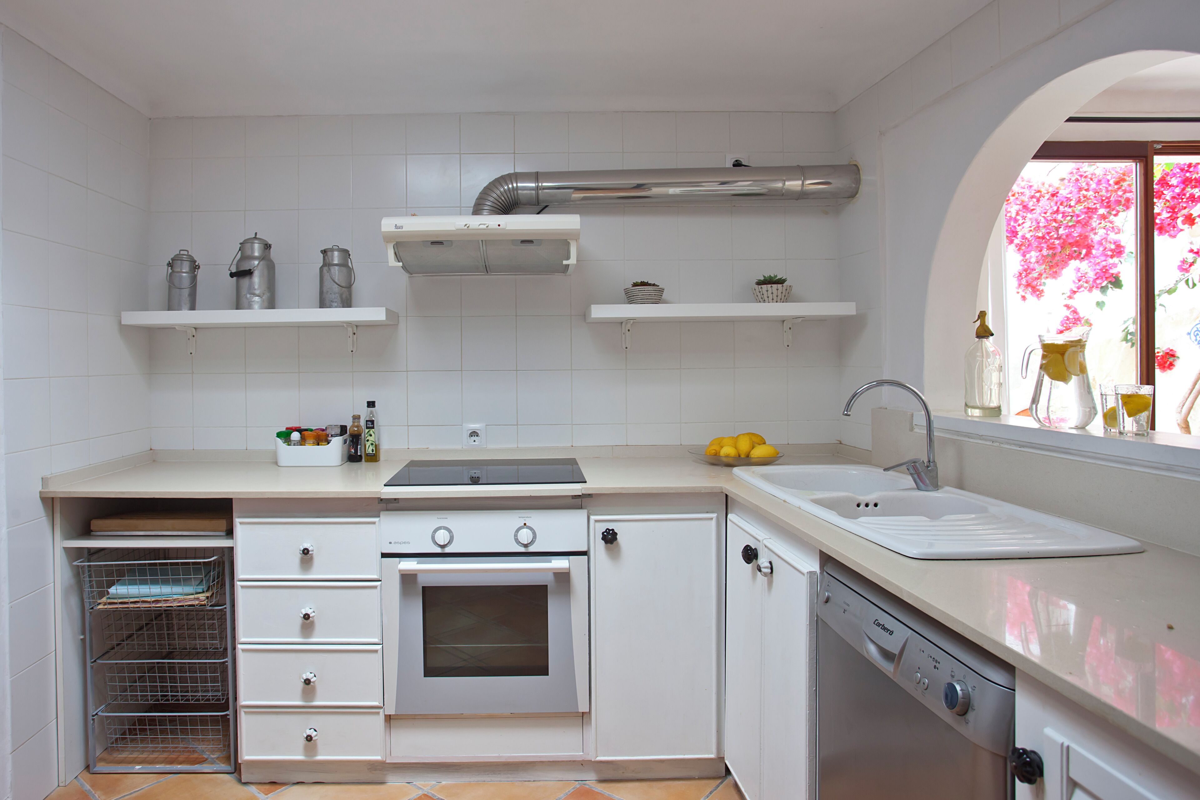 A kitchen with white cabinets, a stove, a dishwasher, and a window with a view of pink flowers.