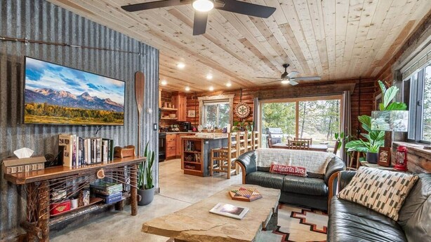 A cozy cabin interior with a wooden ceiling, a large TV mounted on a wall, a dining area with a table and chairs, a sofa with a cushion, and a kitchen area with a grill. - Rustic Trails Near Yellowstone (Fremont County)