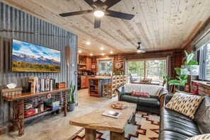 A cozy cabin interior with a wooden ceiling, a large TV mounted on a wall, a dining area with a table and chairs, a sofa with a cushion, and a kitchen area with a grill. - Rustic Trails Near Yellowstone (Fremont County)