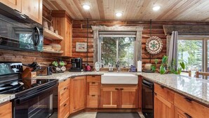 A log cabin kitchen with wooden cabinets, a granite countertop, a built-in stove, a coffee maker, and a window with a view of trees. - Rustic Trails Near Yellowstone (Fremont County)