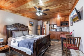 Une chambre comprenant un lit, une table de chevet, un bureau, une chaise, un ventilateur de plafond et un coin cuisine avec des placards et un four à micro-ondes.