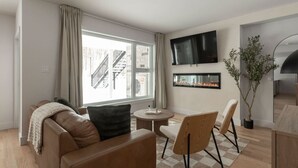 A modern living room with a sofa, chairs, a TV, a fireplace, and a large window with a city view. - Ski In/Out Mont-Blanc | Easy Après-Ski + Fire (Les Laurentides Regional County Municipality)