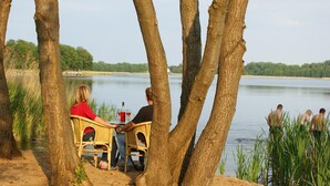A lakeside setting with a dining area under trees, a view of the lake, and people in the water. - Rollstuhlgerechte Ferienwohnung im Ferienpark (Dabel)