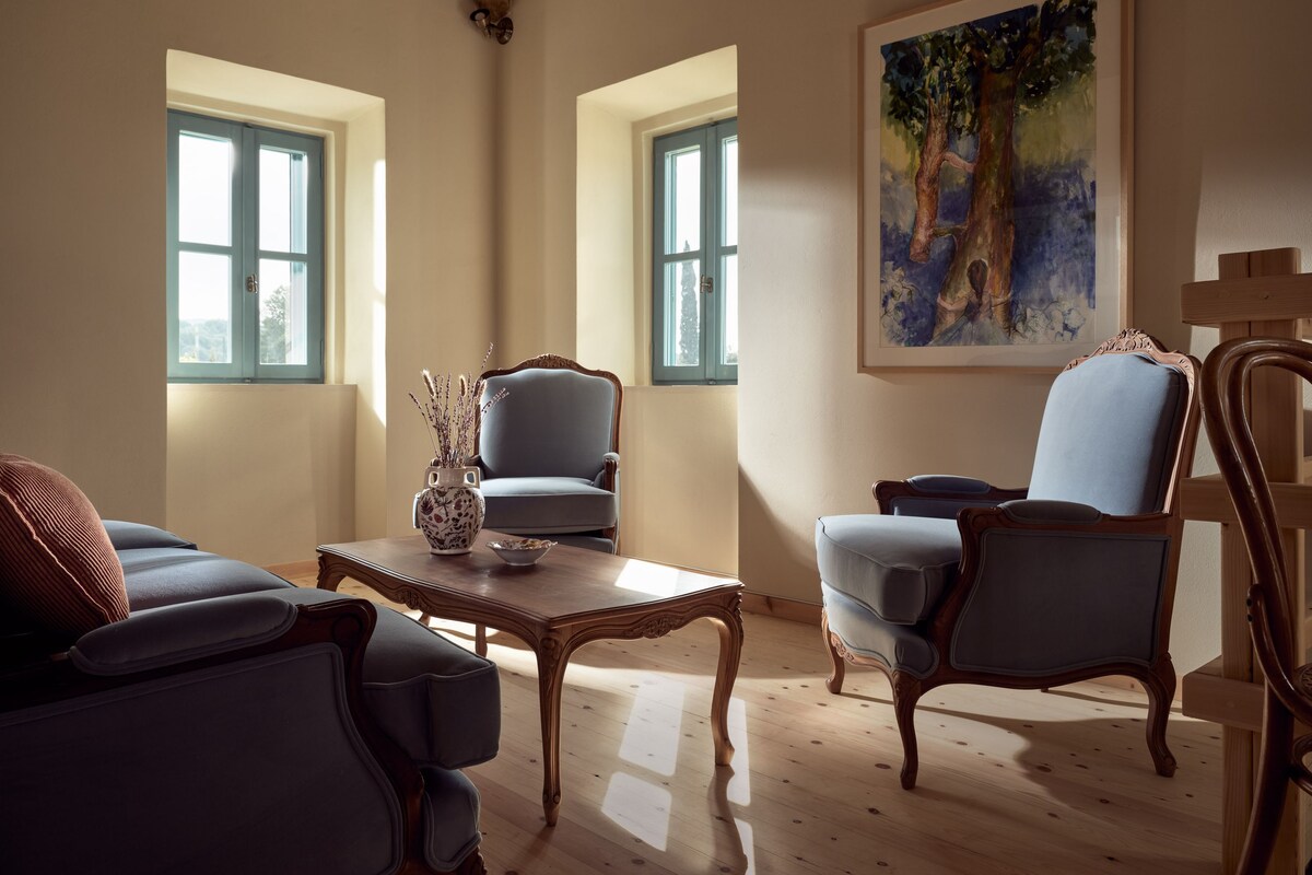 A room with two windows, a painting, a wooden table, and two upholstered chairs.