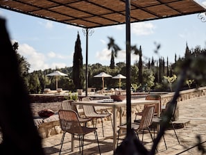 An outdoor patio with wicker chairs and tables, umbrellas, and a view of trees and hills. - Alkis Farm AND Residence (Zakynthos)