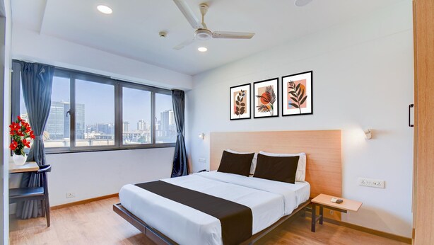 A modern hotel room with a large bed, a desk, a chair, a nightstand, a ceiling fan, and a city view. - Deluxe With Twin Bed Balewadi High (Pune)