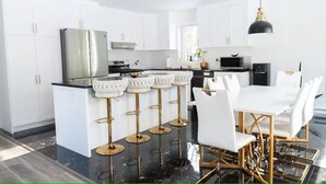 Une cuisine moderne avec des meubles blancs, un îlot central doté de tabourets, une table à manger avec des chaises et un luminaire suspendu.